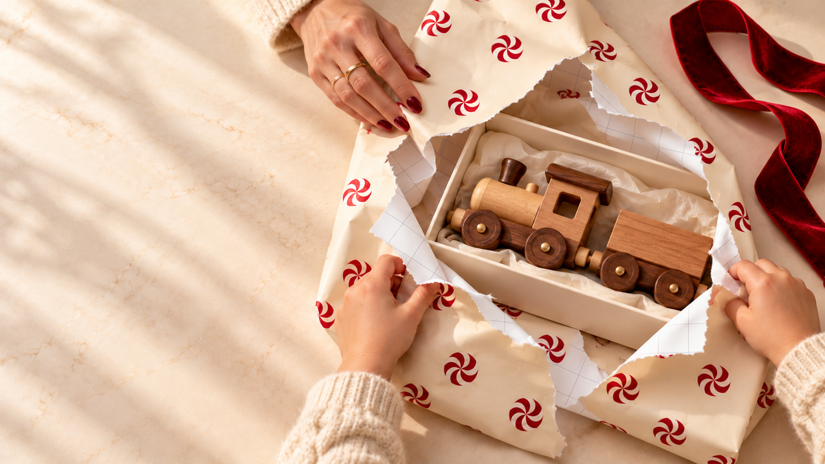 A child opens Maison Velere peppermint gift wrap to reveal a wooden toy train, with cutting grid visible and untied red velvet ribbon beside the gift